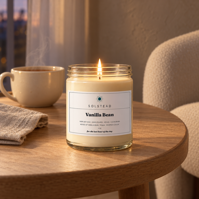 Solstead Vanilla Bean 9 oz soy candle burning on a round wooden side table beside an armchair and a cup of tea