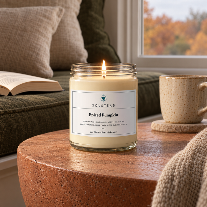 Solstead Spiced Pumpkin 9 oz soy candle burning on a window seat table beside an open book, autumn trees outside